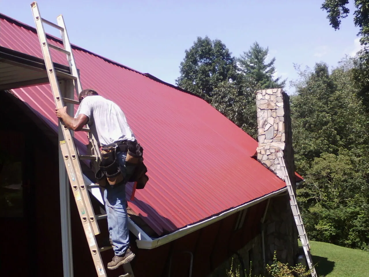 Roofer on a ladder beside a red metal roof and stone chimney — Soffit & Fascia Repair by Roofing Brothers in Fairfax Station